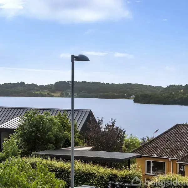 En sø omgivet af grønne bakker med huse i forgrunden, gademast til venstre, blå himmel. Huset til højre har nummer 43.
