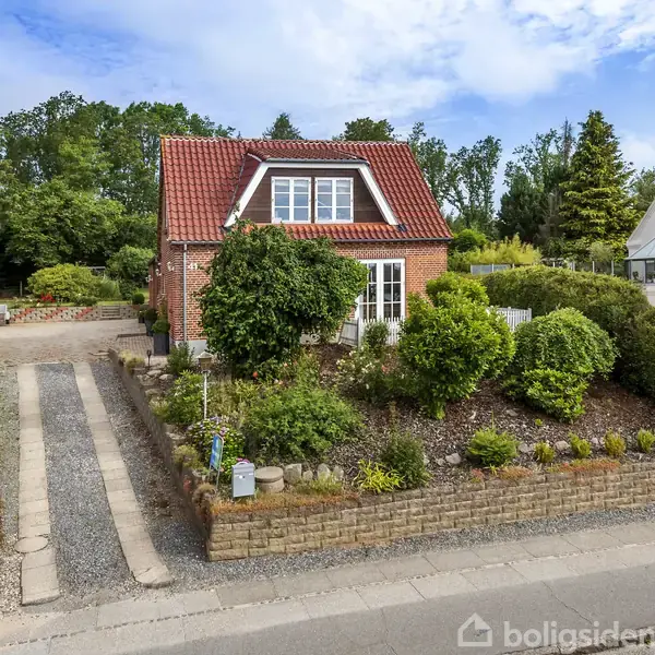 En rødstenshus med rødt tag ligger omgivet af en blomstrende have. Huset har en indkørsel med grus og er omkranset af andre huse og grønne træer i baggrunden.