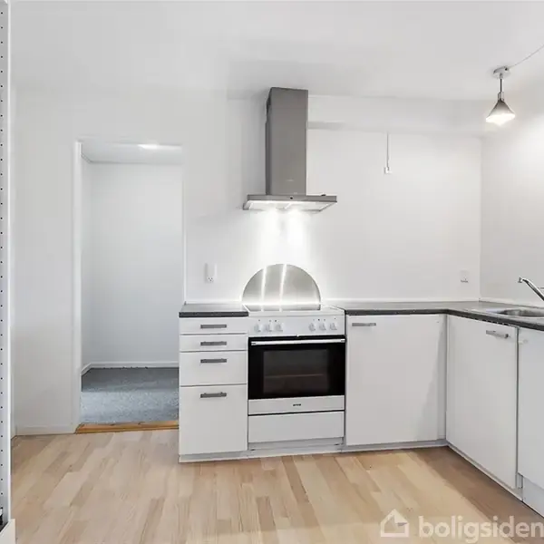 Et moderne køkken har et komfur med emhætte under et skarpt lys, omgivet af hvide skabe og en glat bordplade. Et vindue til højre lader naturligt lys trænge ind.