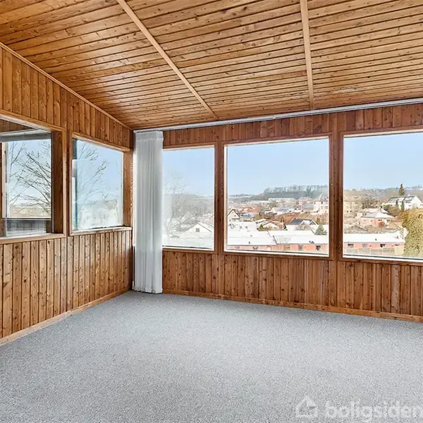Et rum med træpanelvægge og -loft har store vinduer, der giver udsigt over et boligområde med huse og træer. Gulvet er tæppebelagt, og gardiner hænger ved vinduerne.