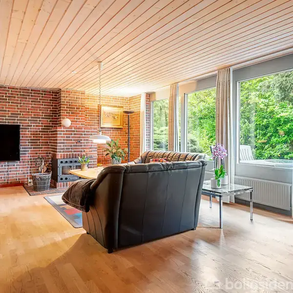 Sort sofa står overfor en væghængt tv-skærm i en stue med trægulv og brændeovn, omgivet af store vinduer med udsigt til en frodig have.