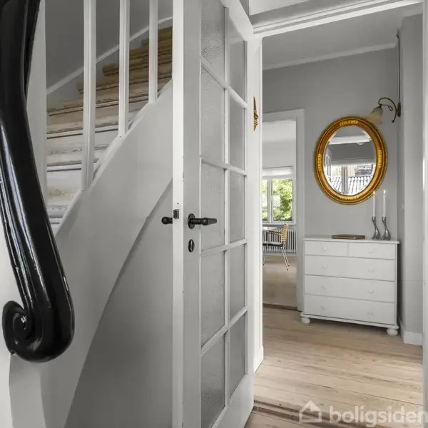 En trappe med mørkt gelænder leder opad ved siden af en åben dør. I baggrunden ses en kommode med et rundt spejl på væggen i et lyst, elegant rum.