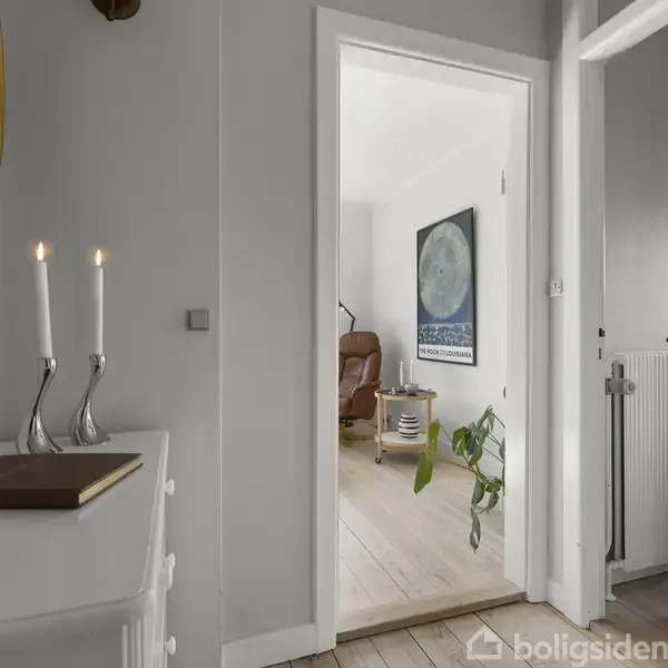 En åben dør fører til en lys stue. I forgrunden ses en hvid kommode med to tændte stearinlys og en bog i en entré med trægulve.