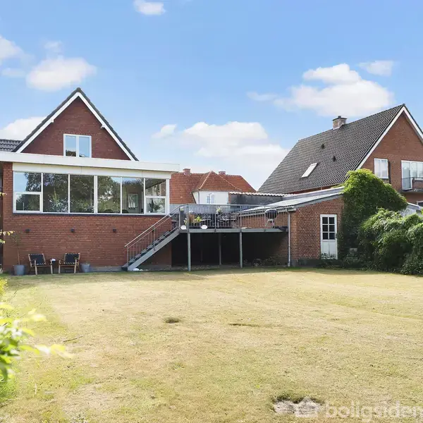 Rød murstensvilla med stor have ses i dagslys. Huset har en høj terrasse med trapper, og omgives af grønne buske og træer. Himmel er blå med få skyer.