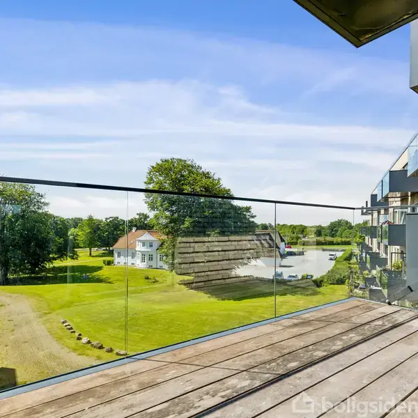 Altan med glasrækværk strækker sig ud fra moderne bygning, tilbyder udsigt over græsplæner, stier og træer. I baggrunden ses et lille, hvidt hus og klart, blåt himmel.
