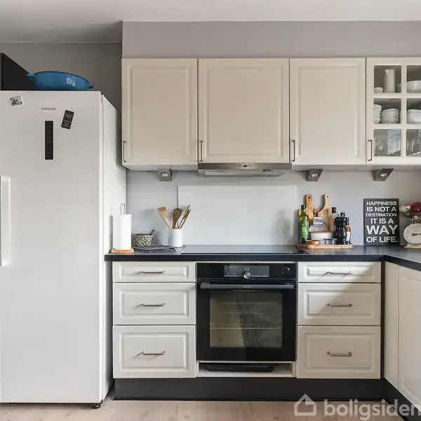 Køleskab står ved siden af et hvidt skab, over en ovn og køkkenbord med madlavningstilbehør. På væggen hænger et skilt: "KITCHEN IS NOT A PLACE, IT IS A WAY OF LIFE".