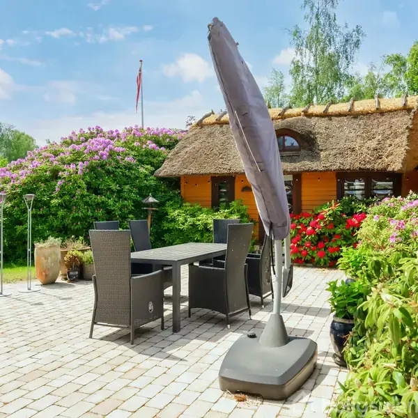 Terrasse med mørk fletmøblergruppe og parasol, omgivet af frodige buske og blomstrende rhododendron; stråtagshus ses i baggrunden, under blå himmel med skyer.