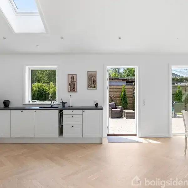 Køkken med hvide skabe og sort bordplade. Sollys strømmer ind gennem vinduerne og en åben dør, der leder til en terrasse med planter. Rundt spisebord ved siden.