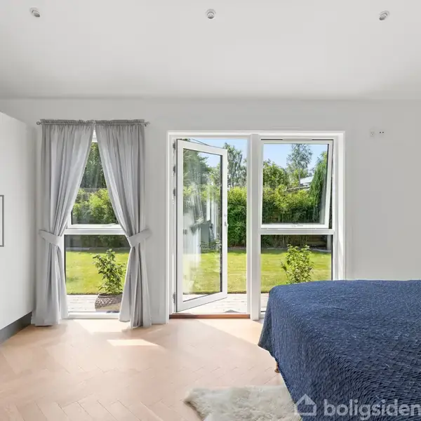 Soveværelse med seng dækket af blåt sengetæppe, skab til venstre, og terrassedør åbner ud mod grøn have med buske. Lyst og minimalistisk indrettet.
