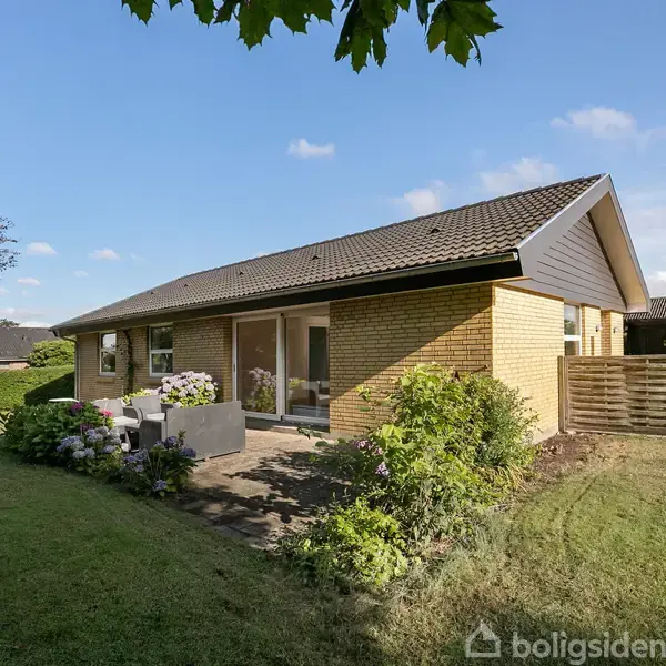 En gul murstensbungalow med sort tag står i en velplejet have med grøn græsplæne og farverige buske. En terrasse med havemøbler ses ved husets side under en blå himmel.