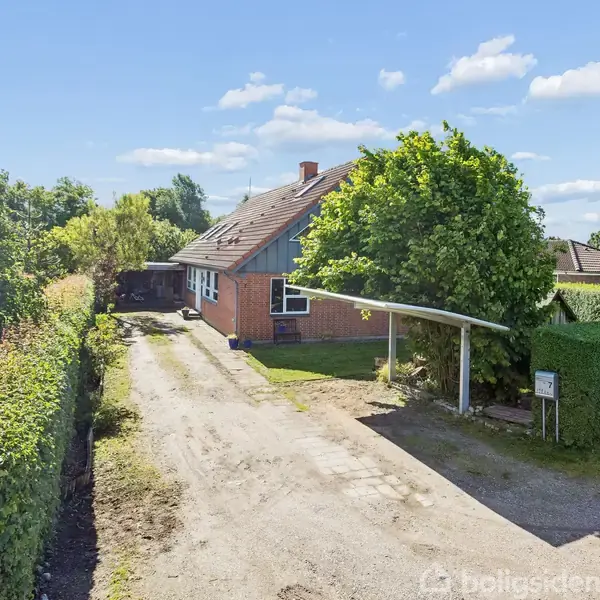 Et rødstenshus står omgivet af grønne hække og en grusvej. I forgrunden ses en carport med træer tæt på. Himlen er blå med enkelte skyer.