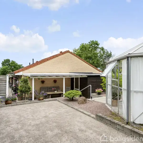 Huset ligger i en gårdhave med grus, omkranset af planters krukker. Til højre ses et drivhus, og bag huset er der grønne træer. Himlen er blå med skyer.