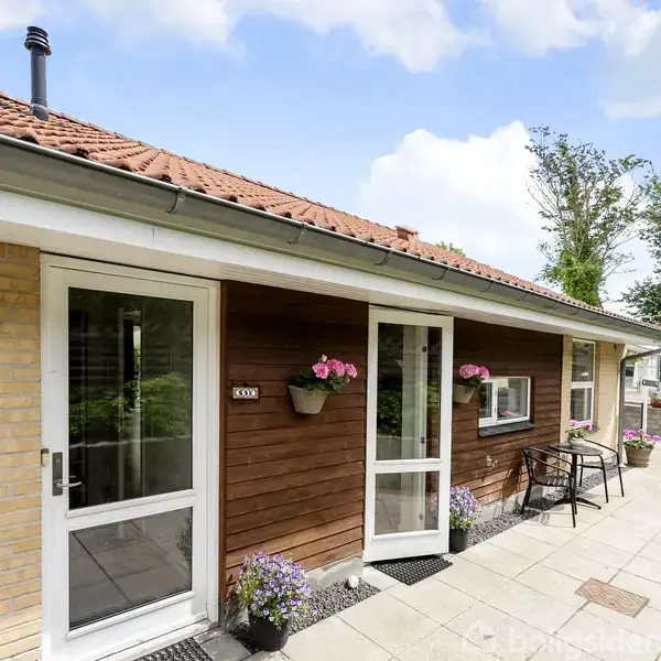 Hus med rødbrune træpaneler og hvide døre. Blomster i krukker langs indgangen. Skilt med teksten "53E" ved siden af døren. Fliser dækker terrassen, træer i baggrunden.