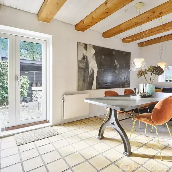 Moderne spisestue med et gråt bord og orange stole omkring det. Store glasdøre fører ud til en terrasse. Træbjælker i loftet og en sort-hvid kunstplakat på væggen.