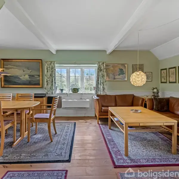 En stue med en brun lædersofa, et sofabord og et spisebord med stole. Rummet har træmøbler, kunst på væggene og er oplyst af naturligt lys fra et stort vindue.