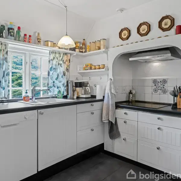 Et lyst køkken med hvide skabe og en sort bordplade. To pendellamper hænger over vasken ved et vindue med blomstrede gardiner. Køkkenredskaber og krydderier er på hylderne.