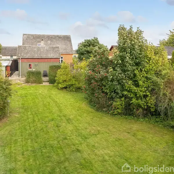 Et lille hus med et gråt tag står i baggrunden. Det er omgivet af en stor, velplejet græsplæne og tæt grøn beplantning, der skaber en frodig have.