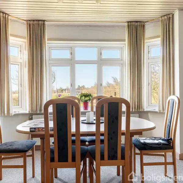Et spisebord med stole står foran et karnapvindue med udsigt til en have. På bordet er en plante og et par genstande. Væggene har dekorationer, herunder et billede.