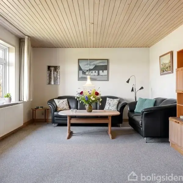 Stue med to sorte sofaer om et sofabord, der har en vase med blomster. Væggene har kunst, og en åben reol står til højre. Store vinduer giver lysindfald.