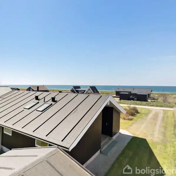 Et sommerhus med mørkt tag og flere små skorstene ses tæt på en kystlinje. Huset er omgivet af græs og flere andre huse. I baggrunden er havet synligt.