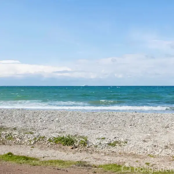 Strand med små sten strækker sig mod det blå og grønne hav, hvor bølgerne ruller ind under en klar, blå himmel med spredte skyer.