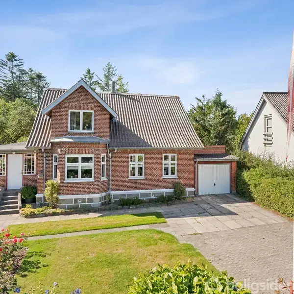 Rødt murstenshus med grå tagsten står i en velholdt have. En flisebelagt indkørsel fører til en garage, omgivet af buske. Flagstang uden flag i forhaven.