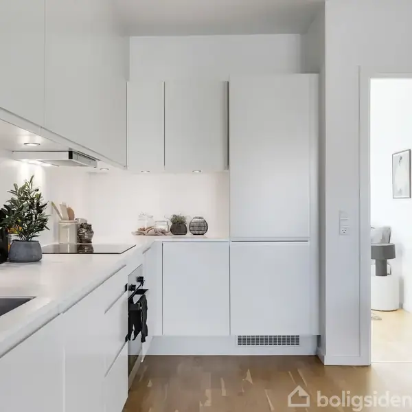 Køkken med hvide skabe og sort bordplade, dekoreret med planter og køkkenredskaber. Indgangen fører til et lyst rum. Trægulv giver varme til det minimalistiske design.