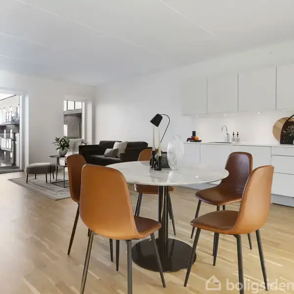 Et rundt spisebord med fire brune stole står i et moderne køkkenalrum med hvidt køkken og trægulve. I baggrunden ses en sofa og en stor stueplante nær vinduerne.