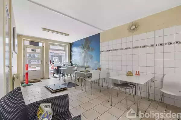 Rummeligt cafeteria med lyse fliser og borde, hvor stole er placeret. Et vægmaleri af palmetræer pryder væggen. Dør til venstre fører ud til gaden. Skiltet siger "ÅBEN".