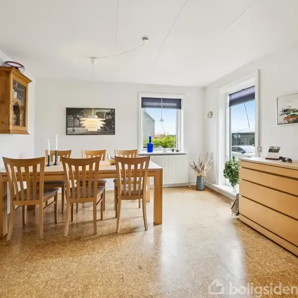 Spisebord med seks træstole står i en lys spisestue. To vinduer giver naturligt lys. Billedet viser hvide vægge, en kommode med lysestager og et væghængt skab.