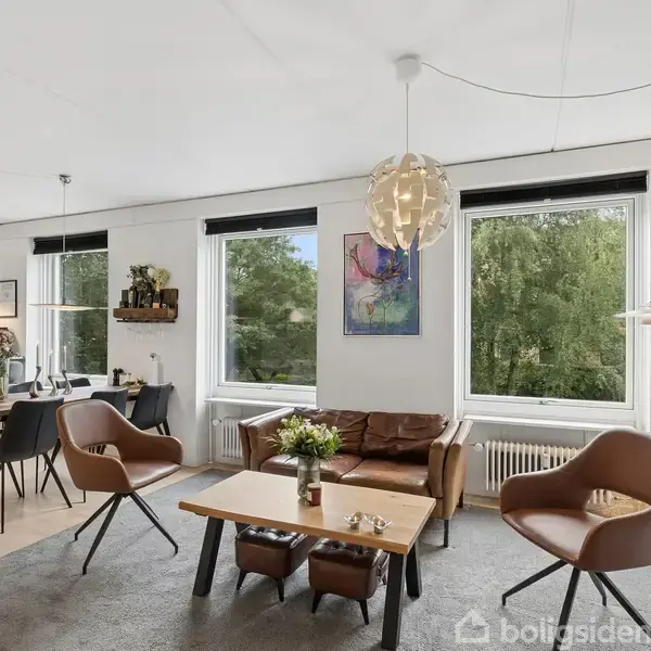 Stue med moderne møbler; to lænestole og en sofa omkring et sofabord. Store vinduer slipper naturligt lys ind. Ved siden er spisestue med bord og stole samt åben køkkenudsigt. Malerier pryder væggene.