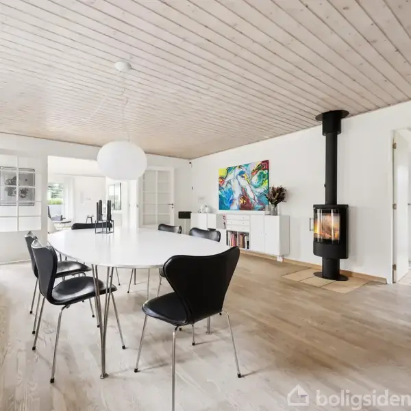 En lys spisestue med et rundt bord og sorte stole. En moderne brændeovn står i hjørnet. Væggene prydes af kunst, og rummet har trægulve og træloft.