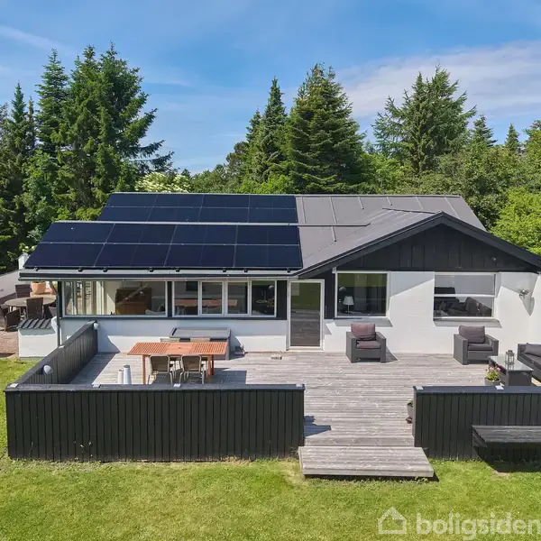 Et moderne hus med solpaneler på taget står i en grøn have omgivet af træer. En stor terrasse foran har udendørs møbler og et spisebord.