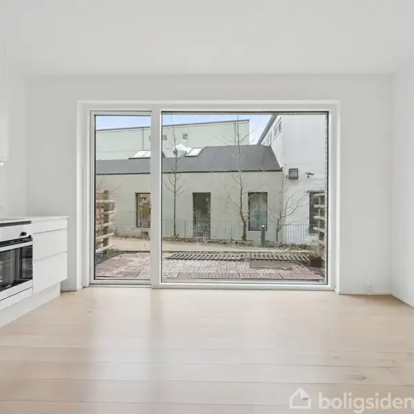 Et hvidt, moderne køkken i et lyst rum med trægulve. Stort vindue med udsigt til en gårdhave med fliser og bygninger. Ingen tekst i billedet.