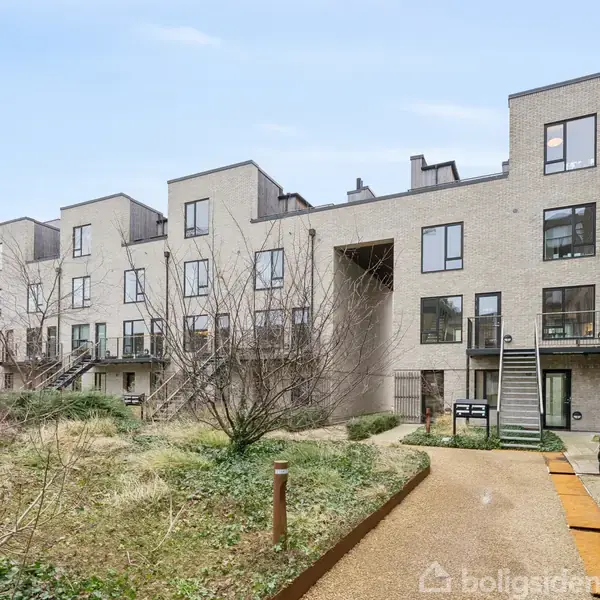 En række moderne, lysegrå boligbygninger står i et åbent område med buske og stier. Bygningerne har store vinduer og altaner. En ladcykel er parkeret i forgrunden.