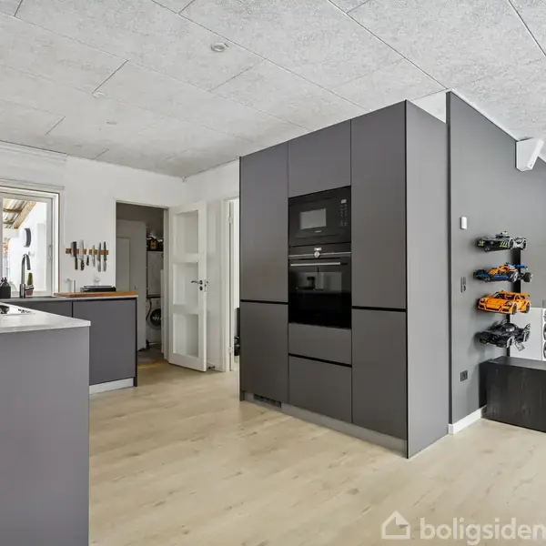 Hvidt hus stående stille i et snedækket landskab, omgivet af buske og træer, med en blå himmel som baggrund.