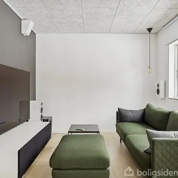 En stue med en stor fladskærm placeret på en lav skænk. Der er en grøn sofa og puf overfor, omgivet af højttalere. Et stort vindue viser træer udenfor.