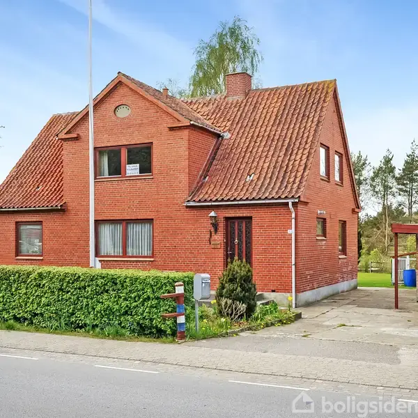 Rødt murstenshus med rødt tegltag står stille omkranset af en hæk i et forstadsområde med træer i baggrunden. Et skur er placeret ved siden af huset.