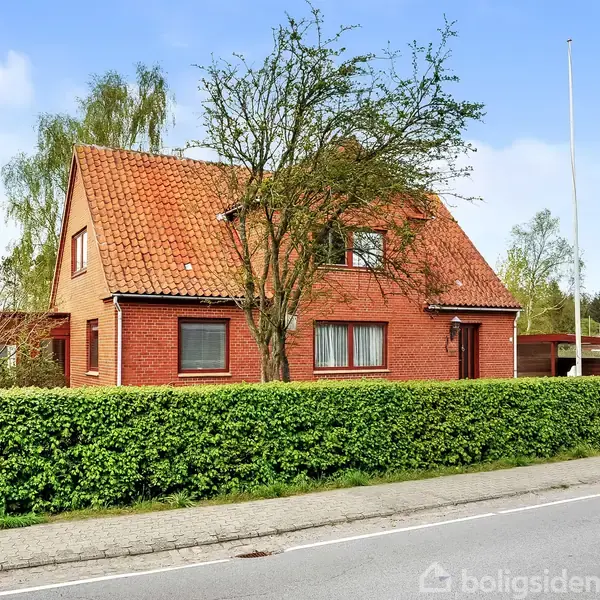Et rødt murstenshus med rødt tag står bag en hæk. Huset har flere vinduer og en dør. Det er omgivet af træer og buske, beliggende ved en vej.