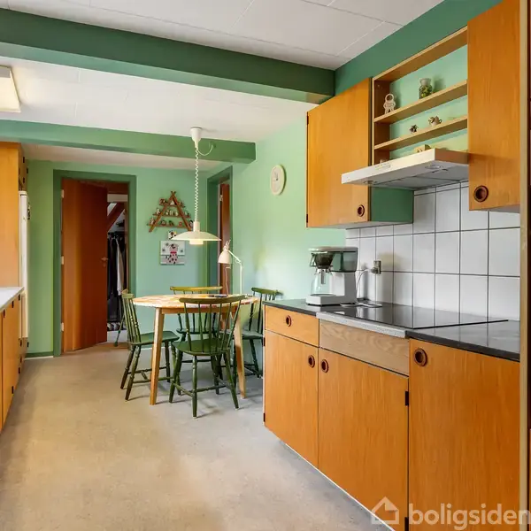 Et køkken med træskabe og hvide fliser over køkkenbordet. I baggrunden står et rundt, træbord med grønne stole. Væggene er lysegrønne, og der hænger en lampe over bordet.