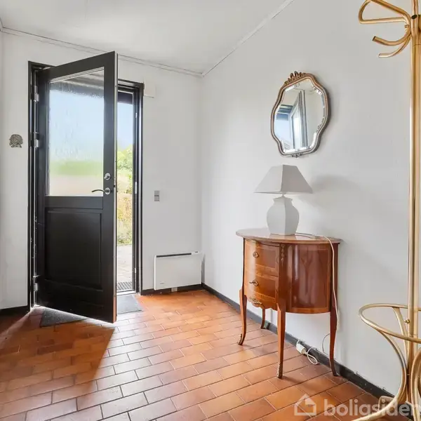 En entré med åben dør, hvor lys strømmer ind. Til højre står et spejl over et lille træbord med lampe. Gulvet er dækket af røde fliser.