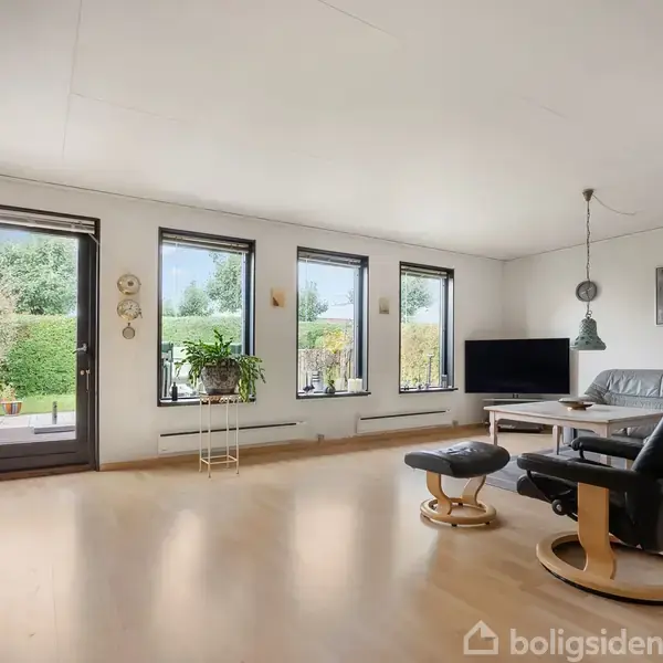En stue med moderne indretning har en lænestol ved en stor fladskærms-TV. Bagved er der store vinduer med udsigt til en have. Der er lyse trægulve og minimalistisk dekoration.