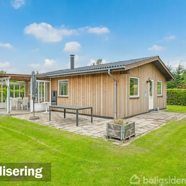 Et lille træhus står på en grøn græsplæne. Til venstre er der en udestue med glasvægge. Teksten "Visualisering" ses nederst til venstre.