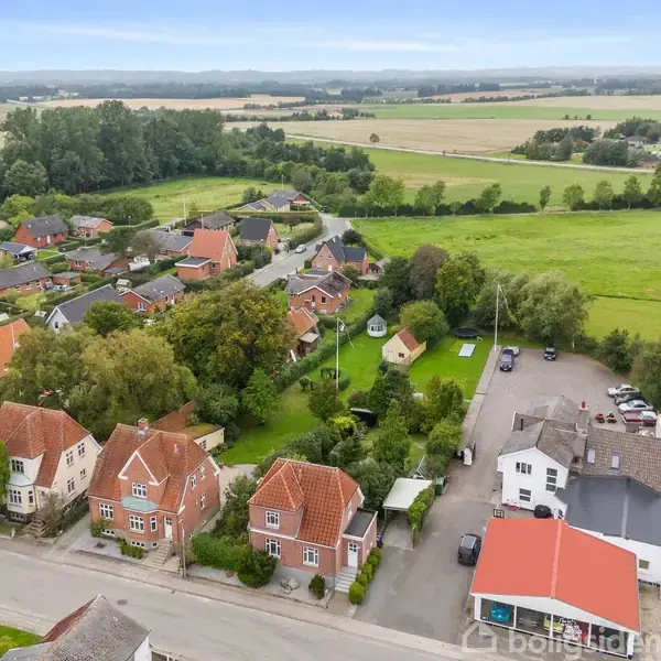 Flere huse med røde tage står langs en vej omgivet af frodige træer og grønne marker i et landligt kvarter med spredt bebyggelse i baggrunden.