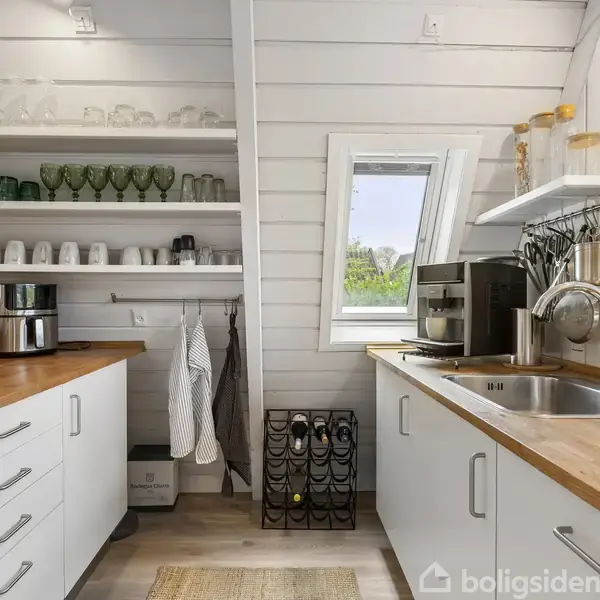 Sort A-formet hus med store vinduer står i en grøn have ved siden af en sø. Flere lignende huse ses i baggrunden, omkranset af træer og buske.