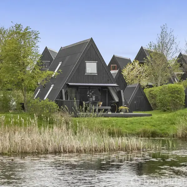 En træterrasse med bænk strækker sig ud mod en sø. Omgivet af grønne plæner og buske, er flere moderne, sorte husfacader synlige i baggrunden under en blå himmel.