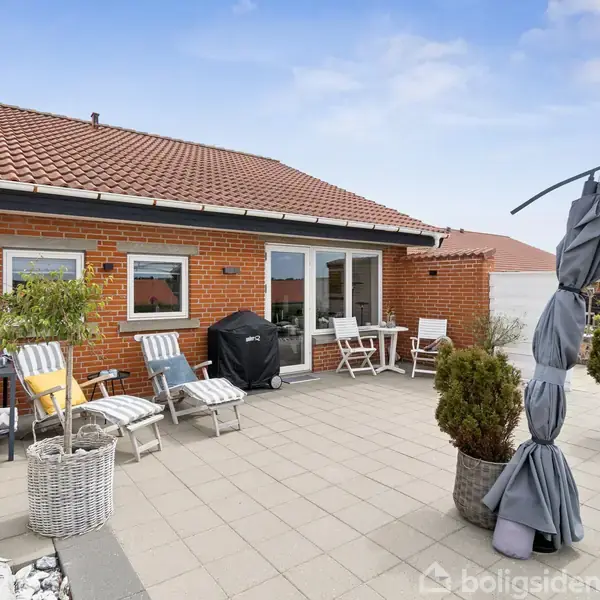 En terrasse med fliser og havemøbler står foran et rødt murstenshus med rødt tegltag. Plantekrukker og en grill er placeret omkring møblerne i en hyggelig gårdhave.