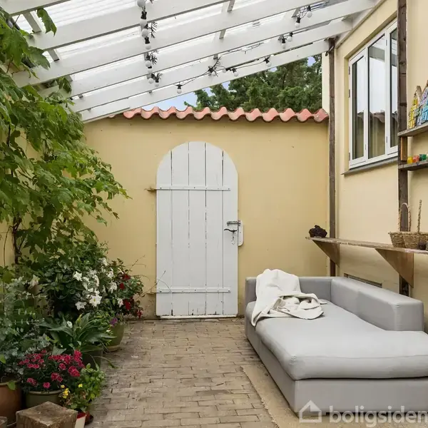 Lys grå sofa står under et transparent tag ved siden af en hvid dør. Omgivet af blomstrende planter og små hylder med dekorative genstande på en væg.