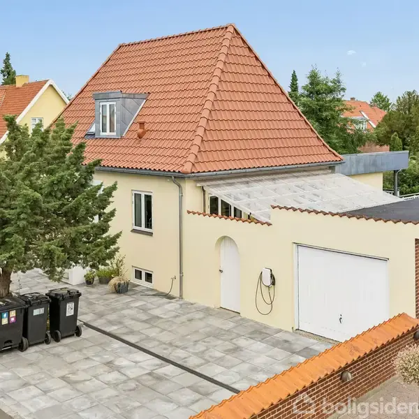 Et hus med rødt tegltag og flere vinduer står på en brostensbelagt indkørsel med affaldscontainere ved siden af et stort træ. Baggrunden viser flere huse og grønne træer.