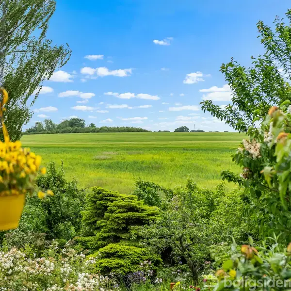 En gul blomsterpotte hænger fra en altankant med en grøn eng i baggrunden. Grønt løv og træer omkranser området under en blå, skyfyldt himmel.